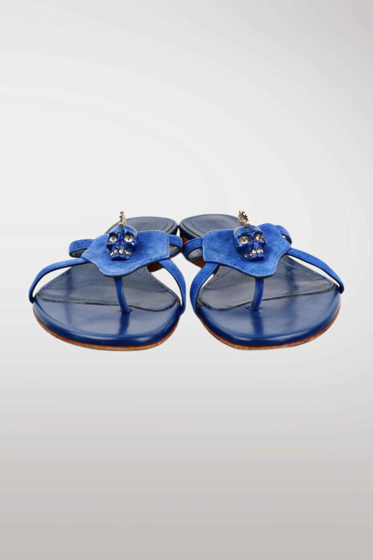 A pair of blue leather thong sandals featuring a sculptural flower-shaped strap adorned with small skull embellishments, set against a light gray gradient background.