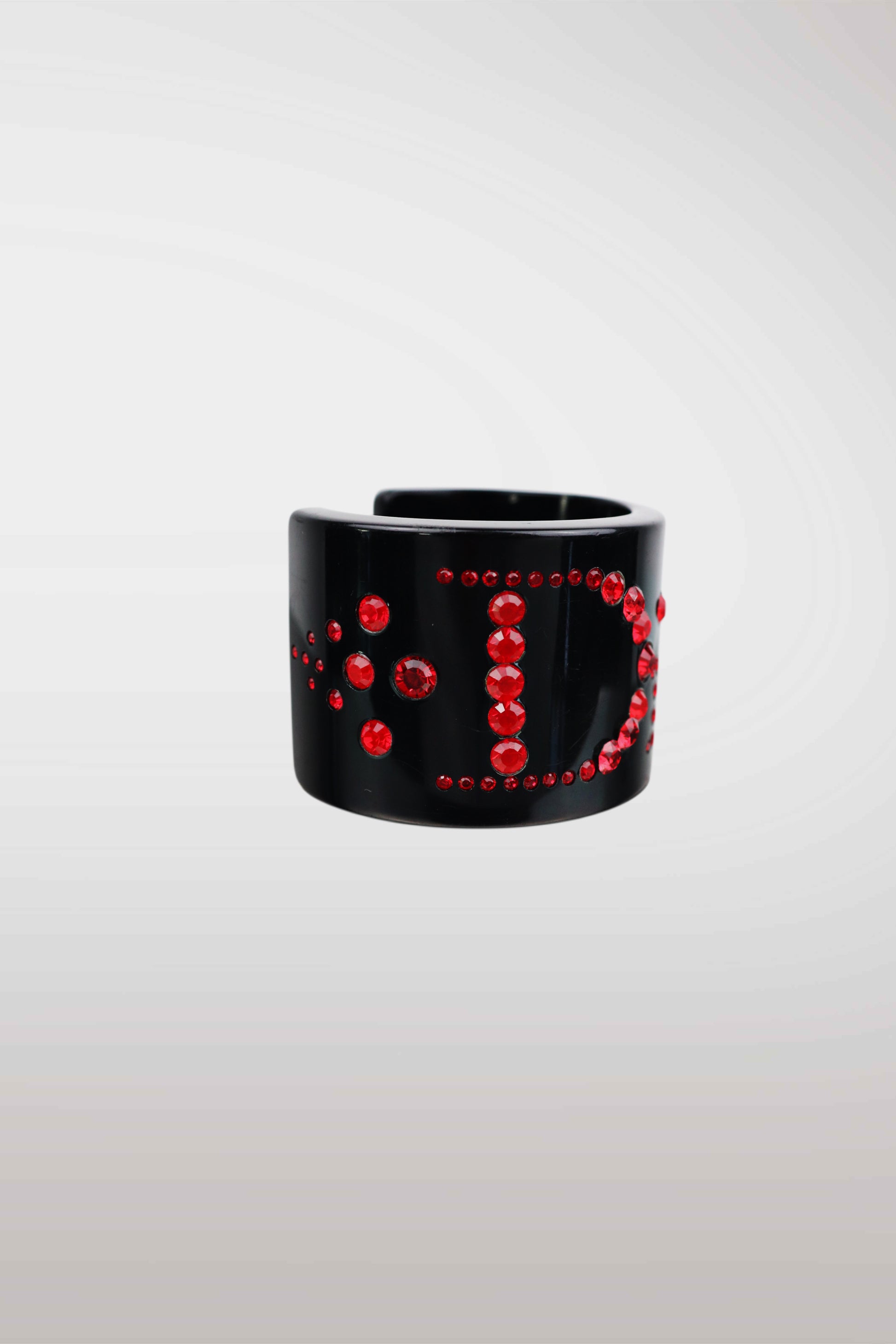 A wide black cuff bracelet embellished with red rhinestones arranged in a circular pattern, shown against a light gray gradient background.