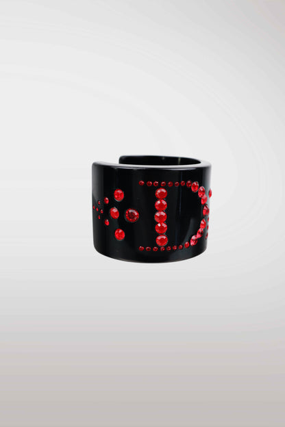 A wide black cuff bracelet embellished with red rhinestones arranged in a circular pattern, shown against a light gray gradient background.