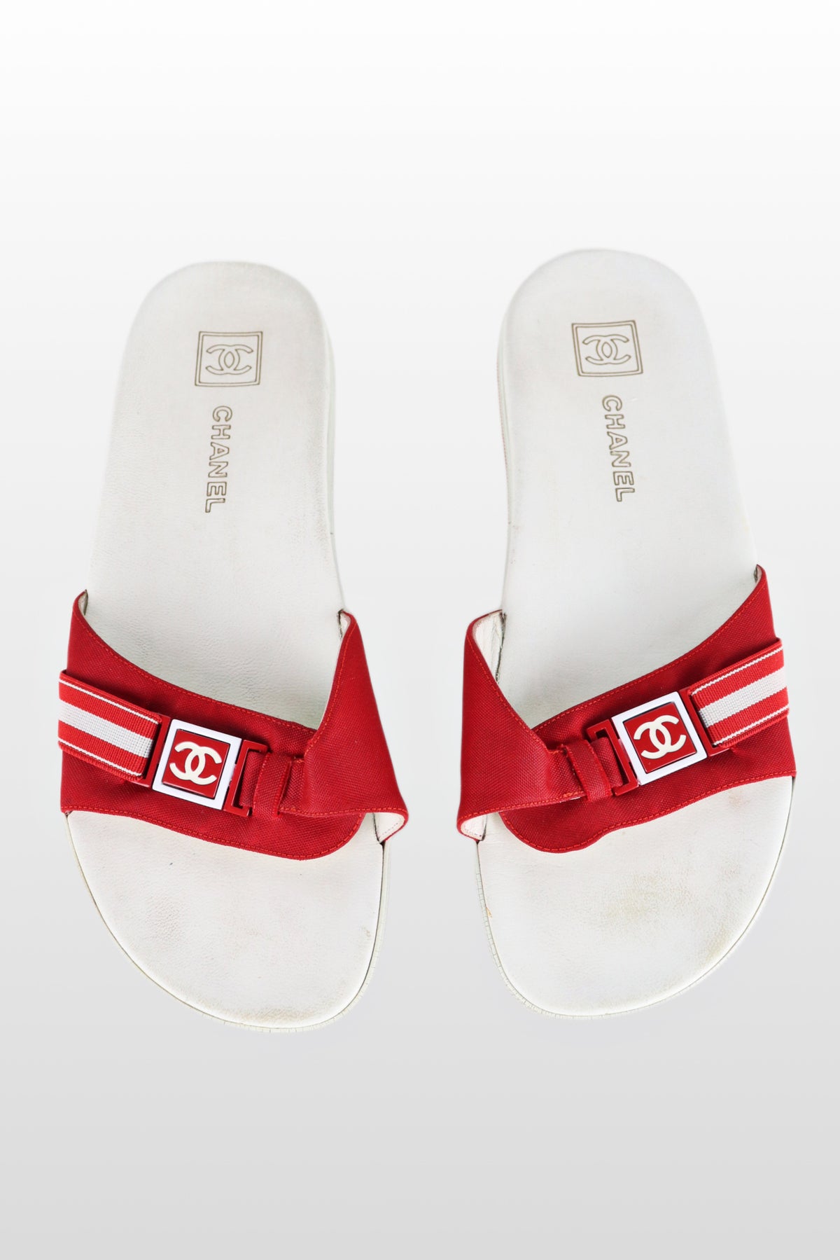 A pair of white leather Chanel sport line slides with a red non-functional buckle detail.