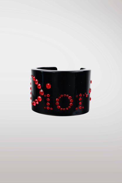 A black cuff bracelet embellished with red rhinestones spelling out 'Dior', shown against a light gray gradient background.