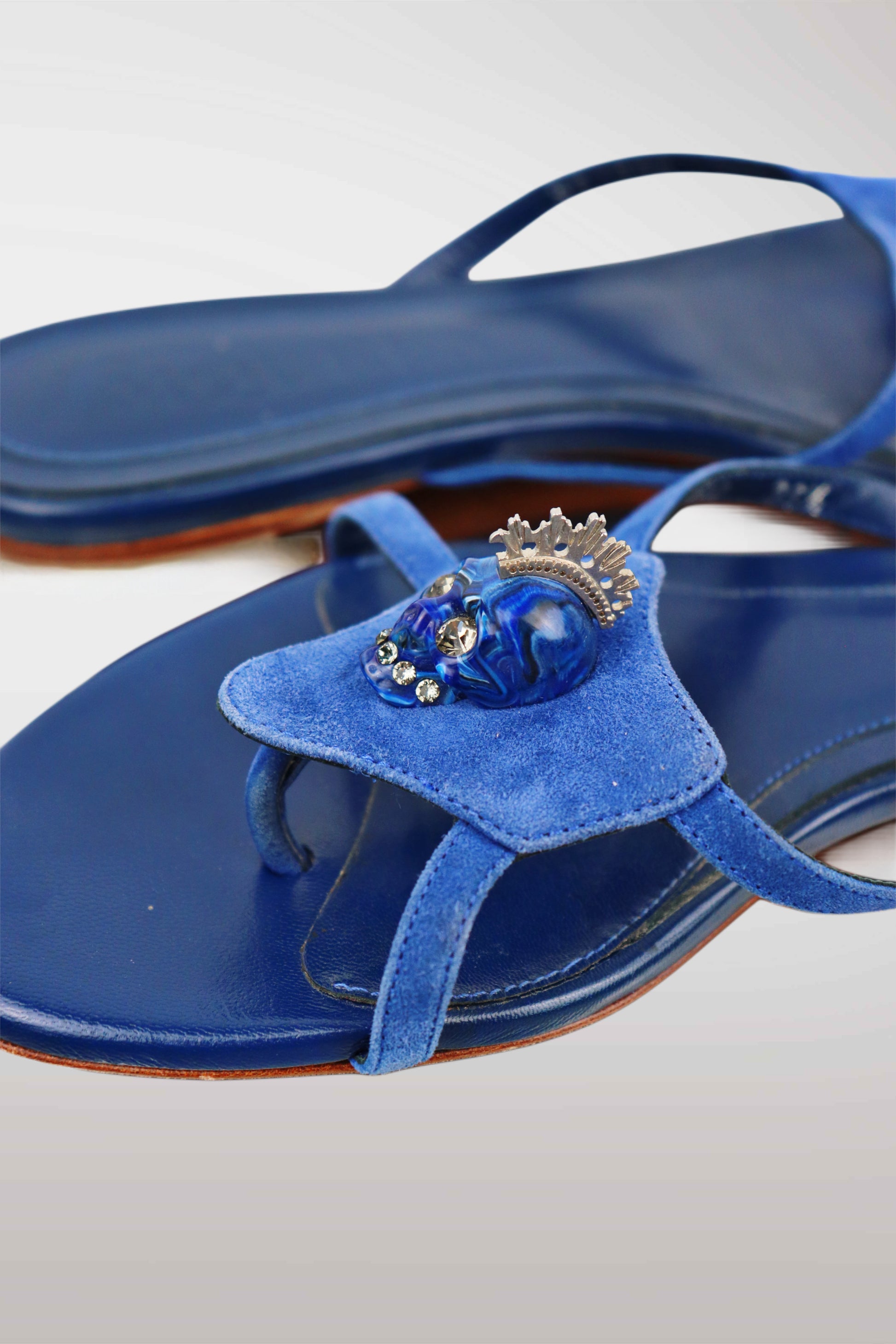 Blue velvet thong sandals featuring a decorative blue skull ornament with a silver crown on the strap, shown on a light gray background.
