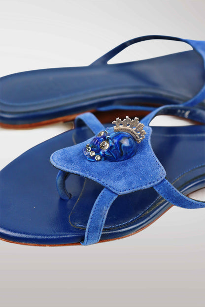 Blue velvet thong sandals featuring a decorative blue skull ornament with a silver crown on the strap, shown on a light gray background.