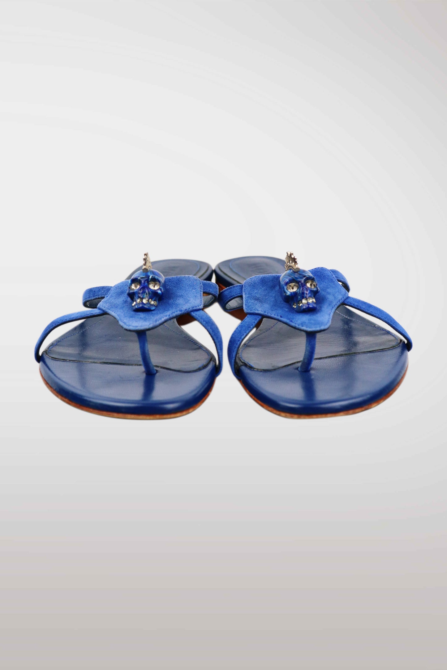 A pair of blue leather thong sandals featuring a sculptural flower-shaped strap adorned with small skull embellishments, set against a light gray gradient background.