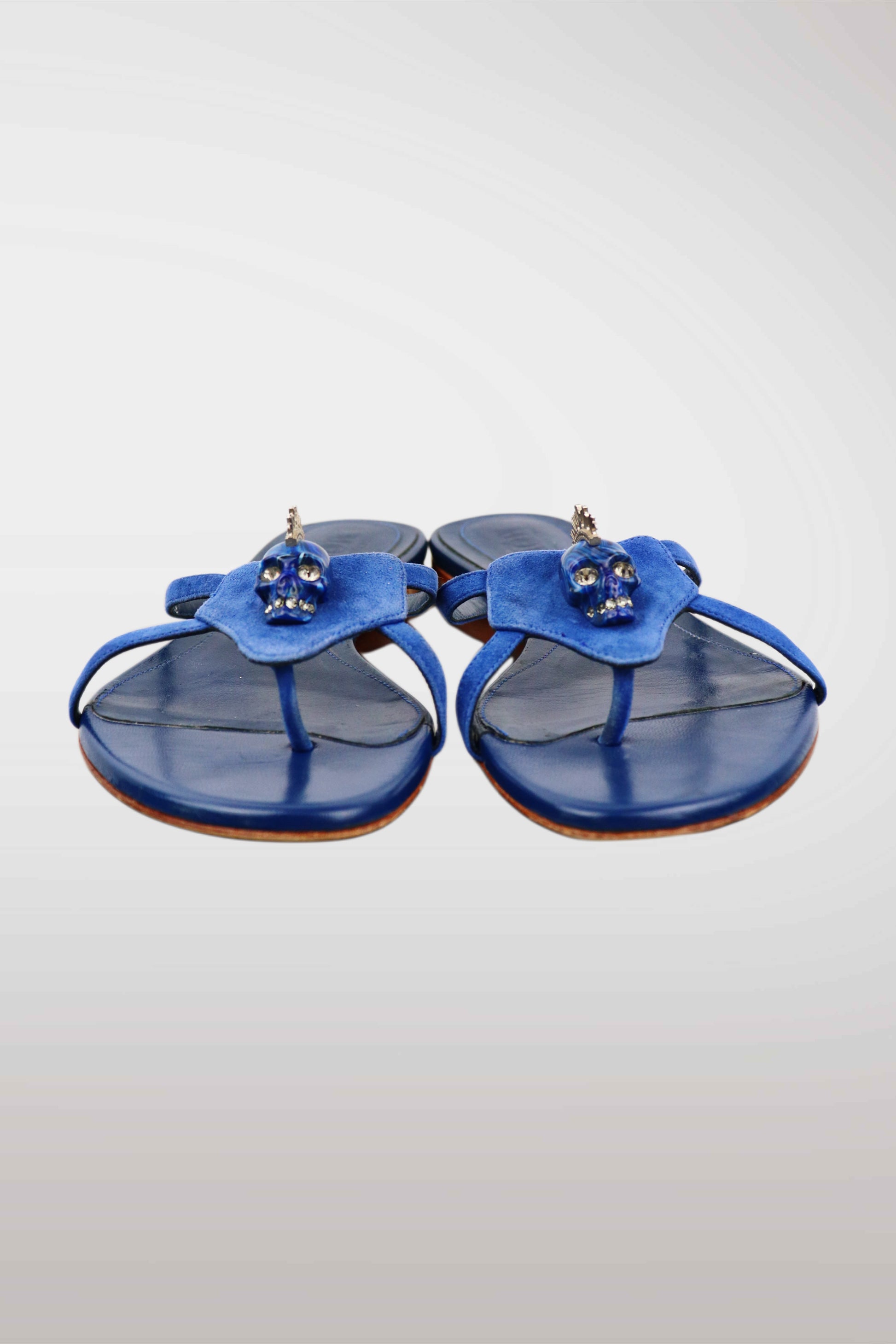 A pair of blue leather thong sandals featuring a sculptural flower-shaped strap adorned with small skull embellishments, set against a light gray gradient background.