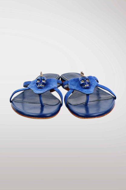 A pair of blue leather thong sandals featuring a sculptural flower-shaped strap adorned with small skull embellishments, set against a light gray gradient background.