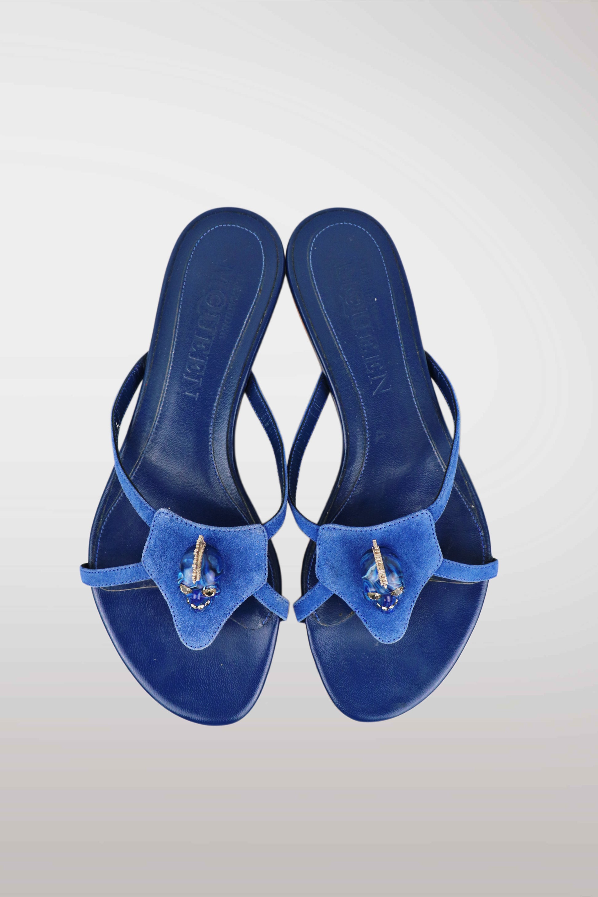 A pair of blue thong sandals with a textured fabric bow detail on the toe strap, embellished with a small gold and blue jewel, shown from a top-down view on a light gray gradient background.
