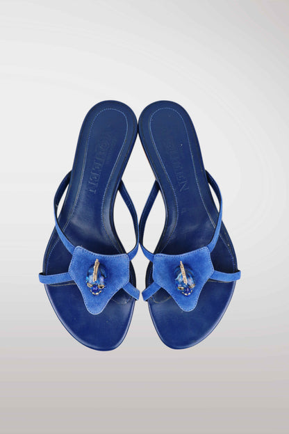 A pair of blue thong sandals with a textured fabric bow detail on the toe strap, embellished with a small gold and blue jewel, shown from a top-down view on a light gray gradient background.