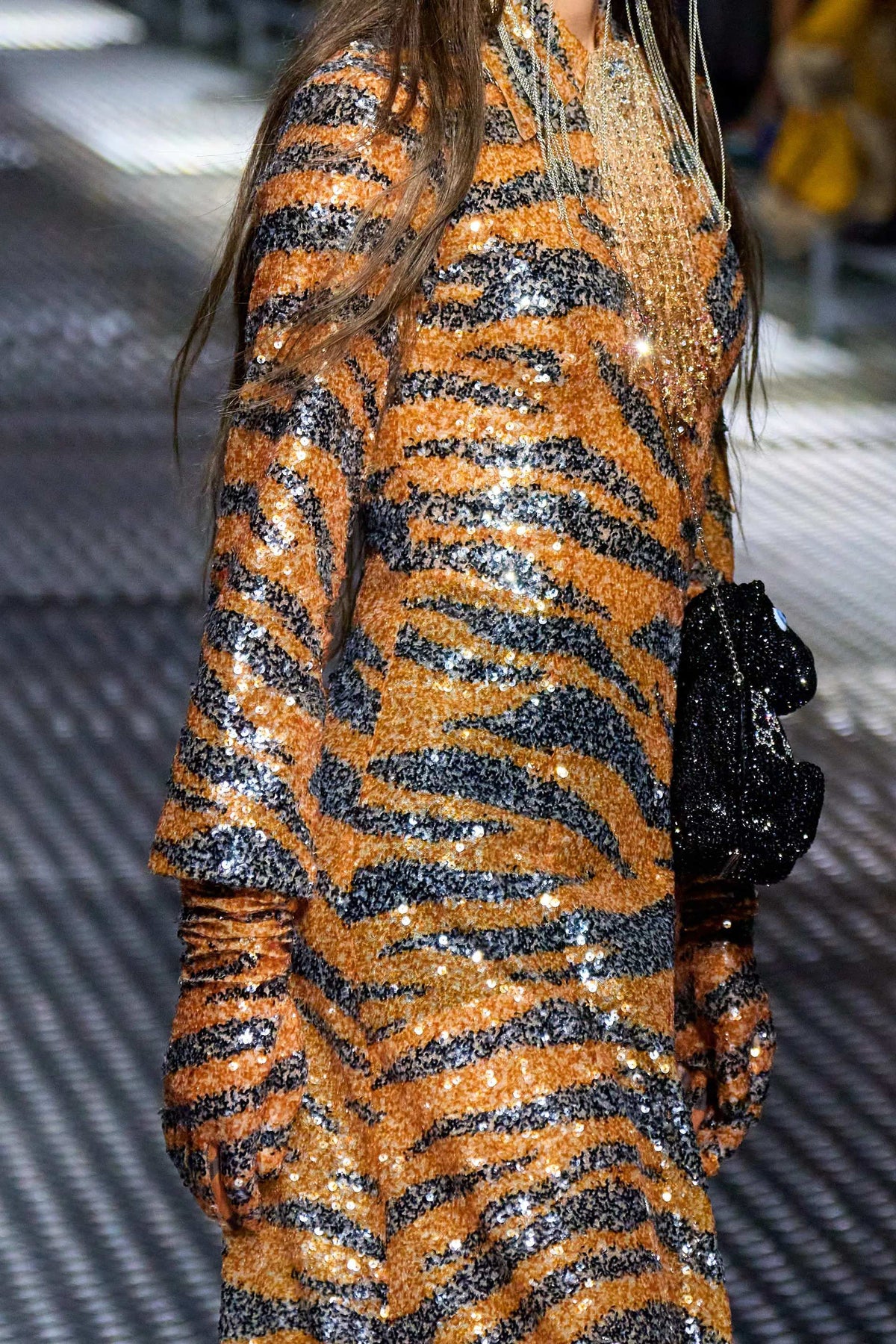 A close-up of a model wearing a glittery orange and black tiger-print sequined dress, holding a black sequined clutch bag.